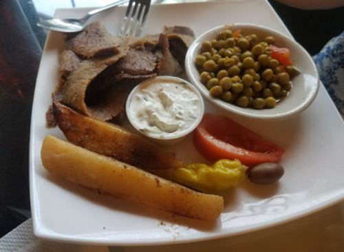 A plate with sliced meat, peas, a tomato slice, a pickled pepper, and a yogurt dip, served with a fork and knife.