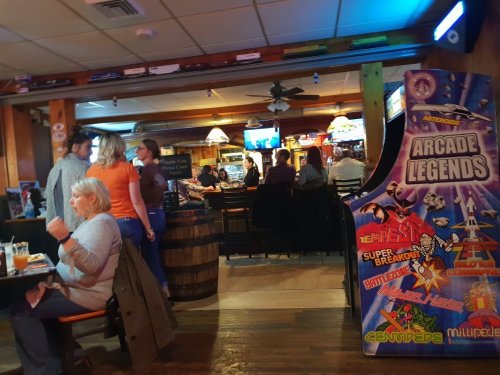 A lively bar scene with patrons socializing, arcade games, and a cozy atmosphere featuring wooden decor.