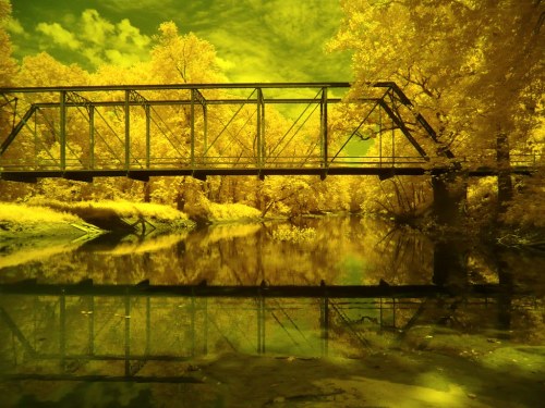 An infrared image of a bridge over a river, surrounded by vibrant yellow foliage and a reflective water surface.