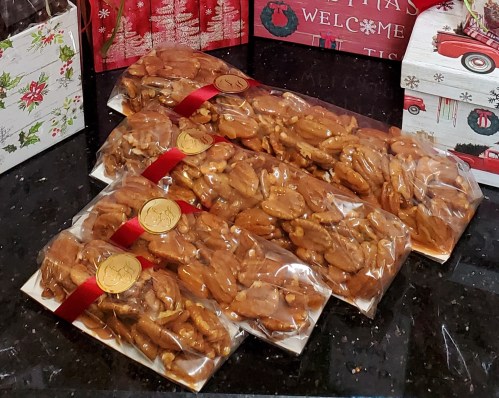 Three bags of candied pecans tied with red ribbons, placed on a dark surface with festive holiday decorations in the background.