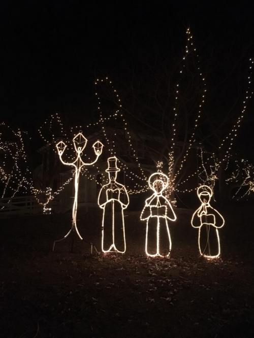 Three illuminated figures in winter attire stand under string lights, surrounded by glowing trees in a festive night scene.