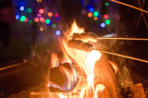 Marshmallows roasting over a vibrant campfire, with colorful lights blurred in the background.