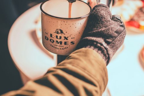 A gloved hand pours a warm beverage from a stylish mug labeled "LUX DOMES" on a table with food in the background.