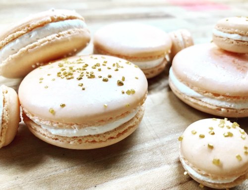 A variety of peach-colored macarons with a creamy filling, some topped with golden sprinkles, on a wooden surface.