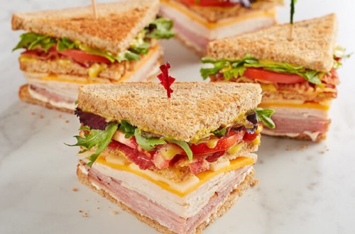 A stacked sandwich with layers of meats, cheese, lettuce, and tomatoes, held together with toothpicks on a marble surface.