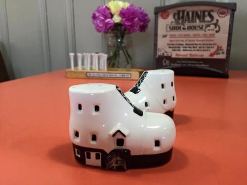 A pair of white ceramic shoe-shaped salt and pepper shakers on a bright orange table, with flowers in the background.