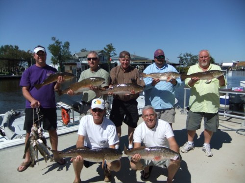 A group of eight men proudly display their catch of fish by a dock on a sunny day.