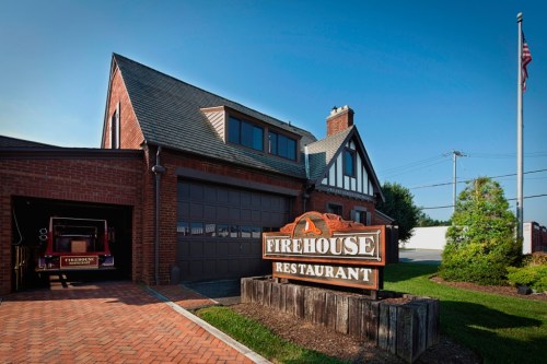 A brick building with a sloped roof and a sign reading "Firehouse Restaurant" in front of a vintage fire truck.