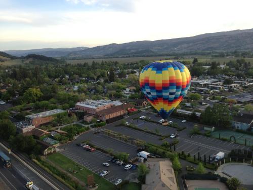 A colorful hot air balloon floats above a small town with green trees and parked cars below.