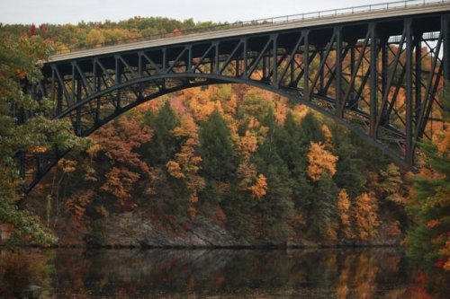 A steel arch bridge spans over a calm river, surrounded by vibrant autumn foliage in shades of orange and green.