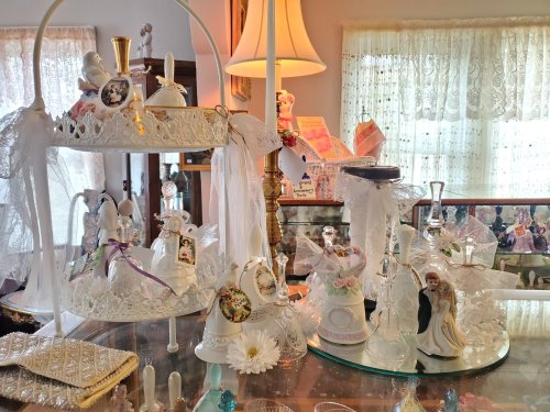 A display of decorative porcelain figurines and bells on a glass table, with lace curtains in the background.