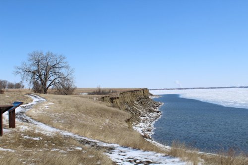 A serene landscape featuring a riverbank, grassy terrain, and a tree under a clear blue sky, with snow on the ground.