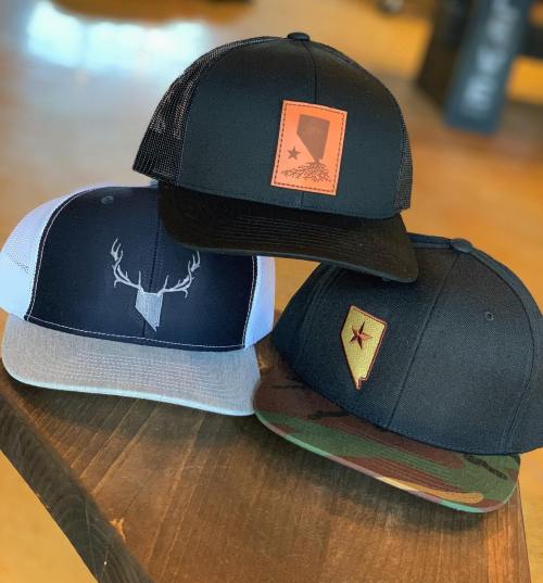 Three stylish hats displayed on a wooden table: one with a deer logo, one with a state emblem, and a camo design.