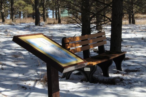 A wooden bench beside a snow-covered area, with an informational sign about the surroundings in a forested setting.