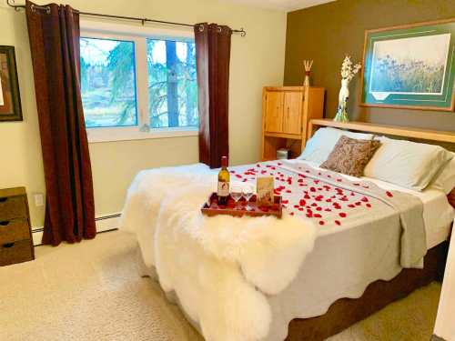 Cozy bedroom with a bed adorned with rose petals, a tray with wine, and a view of trees through a window.