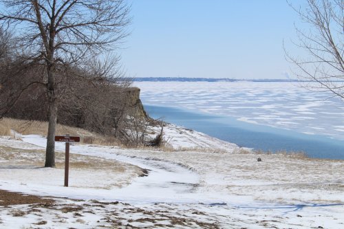 A snowy landscape with a trail sign, overlooking a frozen lake and distant cliffs under a clear blue sky.