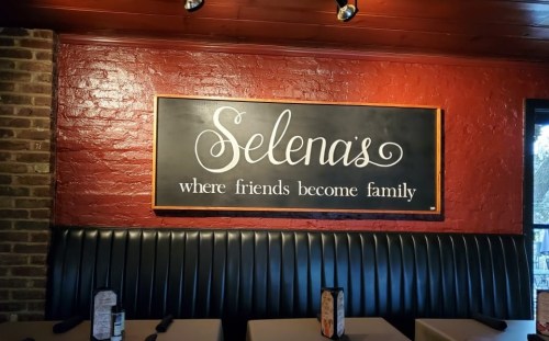 A restaurant interior featuring a sign that reads "Selena's where friends become family" on a red brick wall.