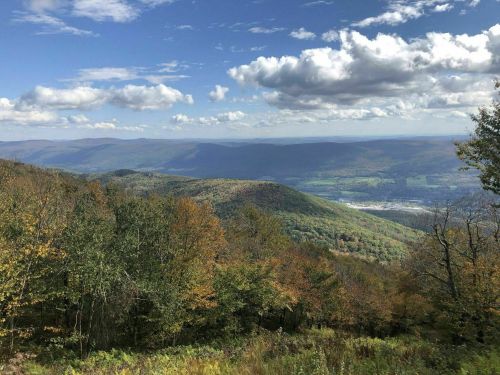 A scenic view of rolling mountains and valleys, adorned with autumn foliage under a partly cloudy sky.