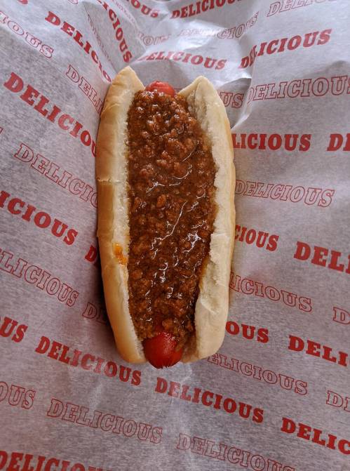 A hot dog in a bun topped with chili, resting on red and white "DELICIOUS" patterned paper.