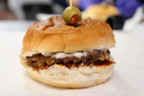 A close-up of a hamburger with a beef patty, mayonnaise, and a green olive on top, served in a soft bun.