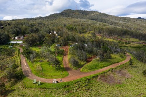 Aerial view of a winding dirt road through lush greenery, with a few buildings and a hill in the background.