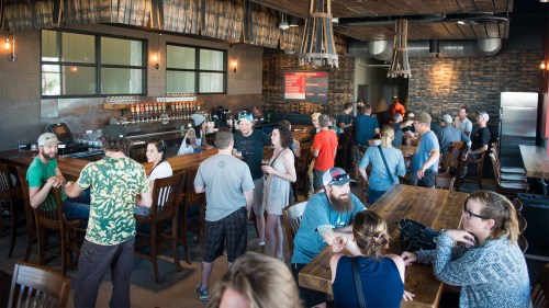 A lively bar scene with patrons socializing, sitting at wooden tables, and enjoying drinks in a rustic setting.