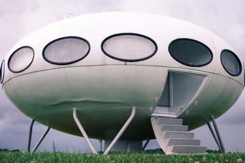A futuristic, white, saucer-shaped structure with round windows and a staircase, set against a cloudy sky.