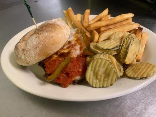 A sandwich with marinara sauce and peppers, served with fries and pickles on a white plate.