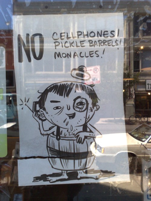 A humorous sign featuring a cartoon character with a phone, stating "No Cellphones! Pickle Barrels! Monocles!"