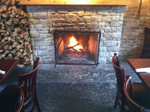 Cozy restaurant interior featuring a stone fireplace with a crackling fire and wooden chairs around tables.
