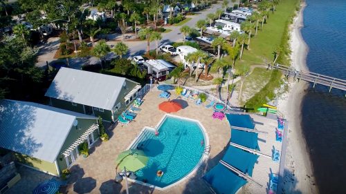 Aerial view of a pool area with lounge chairs, umbrellas, and RVs in the background, surrounded by palm trees.