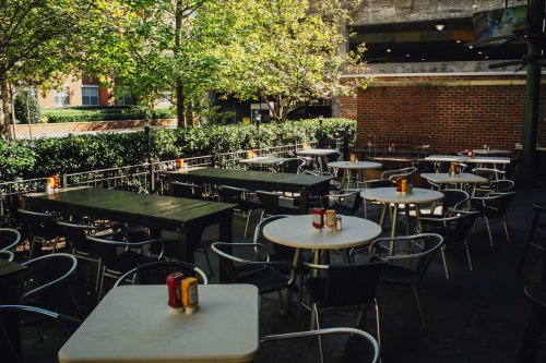 Outdoor dining area with empty tables, surrounded by greenery and brick walls, featuring condiments on each table.