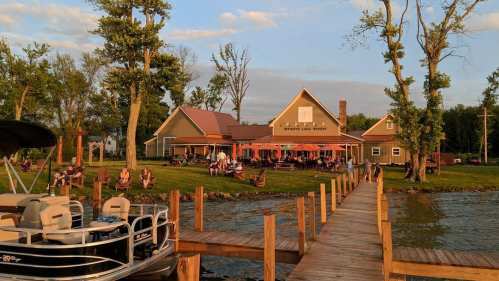 A lakeside winery with outdoor seating, surrounded by trees, and a wooden dock leading to the water at sunset.