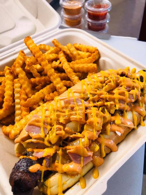 A hot dog topped with mustard and ham, served with crinkle-cut fries and dipping sauces in the background.