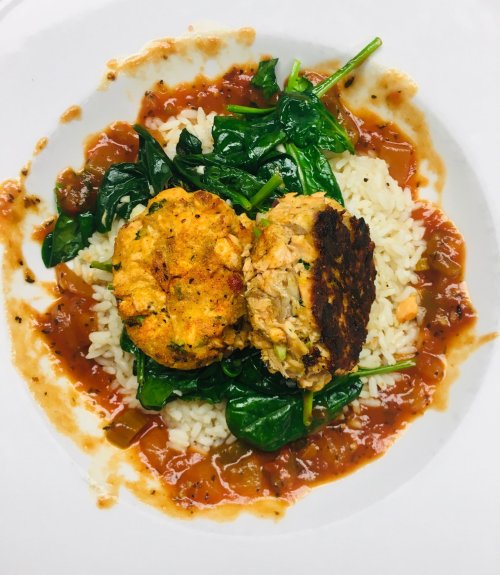 A plate of rice topped with sautéed spinach and two golden-brown crab cakes, drizzled with a rich tomato sauce.