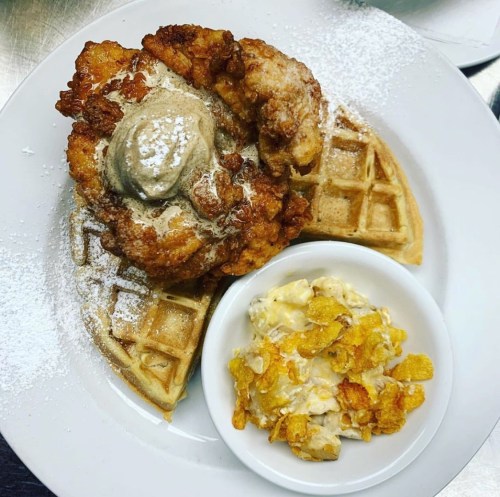 A plate featuring fried chicken on waffles, topped with butter and powdered sugar, alongside a bowl of cornflake cereal.