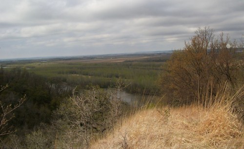 A scenic view from a hilltop overlooking a river and lush green landscape under a cloudy sky.