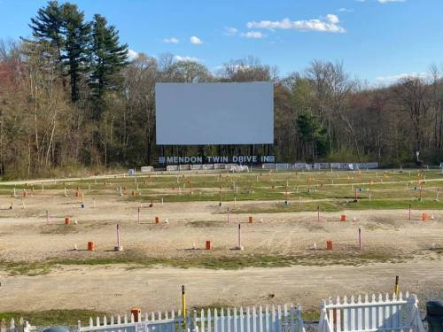 A drive-in movie theater with an empty screen and marked parking spots, surrounded by trees and a clear blue sky.