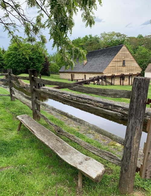 A rustic wooden fence surrounds a grassy area with a bench, leading to a historic building and a small waterway.