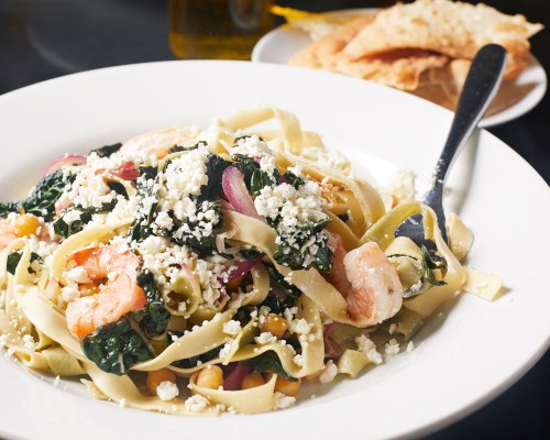 A plate of fettuccine pasta with shrimp, greens, and crumbled cheese, served with a side of crispy bread.