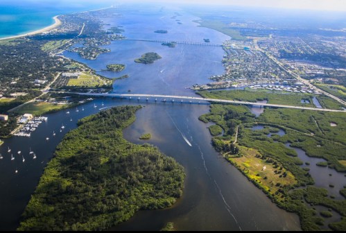 Aerial view of a river winding through lush greenery, with bridges and coastal areas in the background.