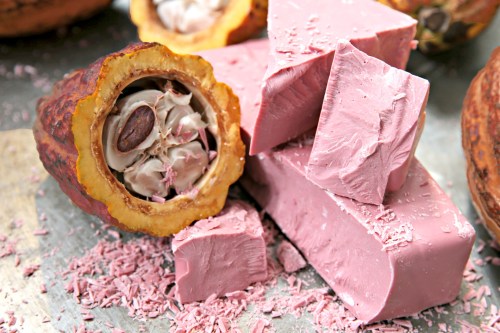 A halved cacao pod reveals seeds next to pink chocolate blocks on a metallic surface.