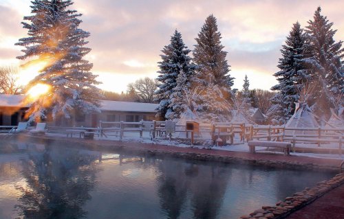 A serene winter scene with a steaming pool, surrounded by snow-covered trees and teepees at sunrise.