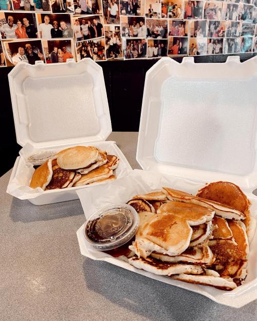 Two takeout containers filled with pancakes, accompanied by syrup, on a table with a collage of photos in the background.