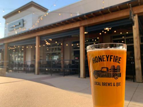 A glass of beer with "Honeyfire Local Brews & Que" logo in front of a restaurant with string lights.