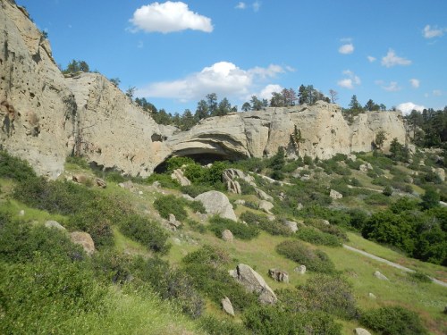 A rocky landscape with a large overhang, surrounded by green vegetation and blue skies with scattered clouds.