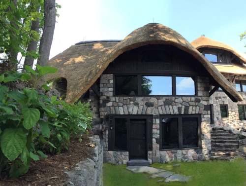 A unique stone house with a thatched roof, surrounded by greenery and featuring large windows.