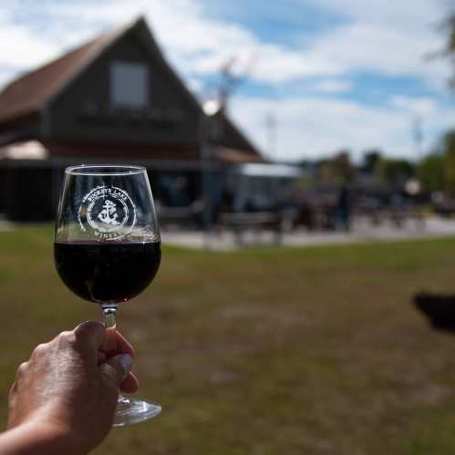 A hand holding a glass of red wine in front of a winery building and outdoor seating area on a sunny day.