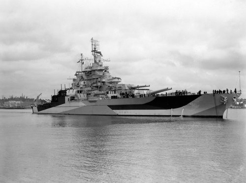 A large battleship docked in calm waters, with crew members visible on deck and a cloudy sky in the background.