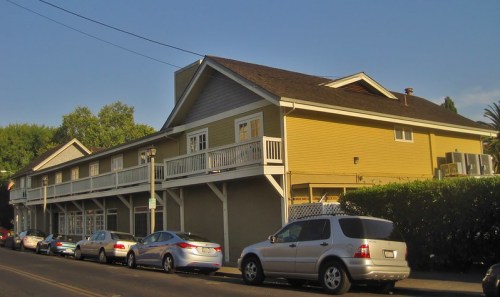 A two-story yellow building with balconies, surrounded by parked cars along a tree-lined street.